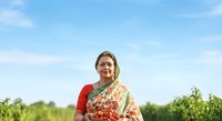 Middle-aged woman of South Asian ethnicity wearing traditional red and gold embroidered sari with dupatta, standing in lush green agricultural field under clear blue sky. Suitable for cultural diversity, farming, rural lifestyle, and traditional clothing concepts.