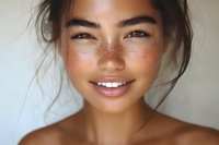Close up portrait of young Asian woman with freckles and natural makeup smiling at camera, showing bright white teeth and glowing skin against light background.