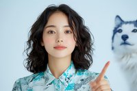 Young Asian woman with wavy black hair wearing turquoise floral pattern shirt pointing upward while husky dog looks on in background against light blue wall.