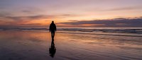 Silhouetted surfer in wetsuit standing on wet beach sand at golden hour sunset, holding surfboard, ocean waves and dramatic sky backdrop for travel and lifestyle content.