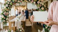 Woman in pink dress holding blank white card at elegant floral wedding venue with guests and decorative flowers in background, suitable for invitations and event announcements.