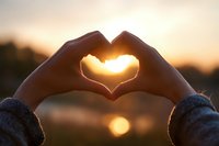 Hands forming heart shape silhouette against golden sunset sky with warm sunlight glowing through fingers creating romantic love symbol for Valentine's Day.