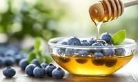 Golden honey dripping from dipper onto fresh blueberries in glass bowl with mint leaves, natural light and soft bokeh background creates appetizing food scene.