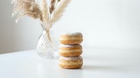 Stack of three golden brown donuts dusted with powdered sugar on white table with dried pampas grass in crystal vase creating minimalist bakery scene.