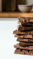 Stacked dark chocolate pieces with textured surface on white background, kitchen setting with blurred bowls and containers behind, suitable for food and culinary design projects.