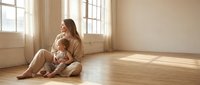 Young Caucasian mother in beige outfit sitting on wooden floor holding toddler son in bright sunlit room with large windows for family lifestyle content.