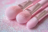 Pink makeup brushes with glitter sparkles scattered on surface, soft focus close-up shot showing professional cosmetic tools with rose gold handles for beauty application.