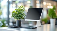 Modern workspace with tablet on stand, wireless keyboard and green plant in pot against blurred office background, emphasizing minimalist home office setup.