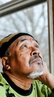 Senior male, East Asian ethnicity, wearing neon yellow-green shirt and black beanie, gazing upward thoughtfully by window with blurred winter branches visible behind glass.