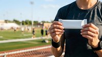 Young adult male athlete holding blank white ticket or pass at sports field stadium during daytime event, suitable for sports marketing and event promotion materials.