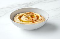 Fresh hummus served in white bowl on marble surface, drizzled with olive oil and paprika, traditional Middle Eastern chickpea dip photographed in natural light.