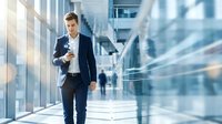 Young professional businessman walking through modern office corridor with coffee cup during busy workday in corporate building.