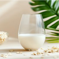 Fresh oat milk in glass with scattered oats on light surface, green palm leaf in background. Natural dairy alternative beverage styling for healthy lifestyle.