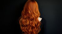 Young woman with long wavy copper red hair shown from behind against dark background, demonstrating luxurious healthy hairstyle with natural-looking curls.
