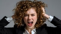 Young woman with curly red hair screaming in frustration while grabbing her head, wearing black blazer and white shirt against gray background.