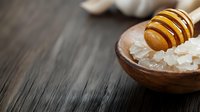 Wooden honey dipper dripping golden honey over sea salt crystals in rustic bowl on dark wooden background, natural spa ingredients for skincare.