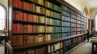 Extensive library shelving displays hundreds of colorful books organized by spine color gradient from red through yellow, green, and blue in an elegant interior space with arched windows and classical architecture.