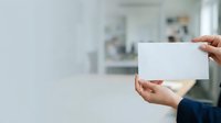 Hand holding blank white business card or paper in modern office environment with blurred background, suitable for branding, corporate identity, and marketing materials.