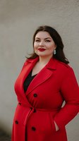 Confident plus size woman in bright red coat and red lipstick smiling against neutral wall background. Professional fashion portrait showcasing body positivity and style.