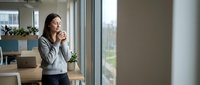 Young professional woman enjoying coffee break in modern office space looking out window during workday pause.
