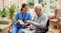 Young female healthcare worker in blue scrubs using tablet with elderly Caucasian man in wheelchair during home care visit in bright living room.
