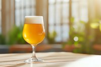 Glass of cold craft beer with foam head on wooden table against blurred window background with warm sunlight and bokeh effect.