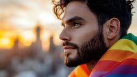 Young Hispanic man with beard wearing rainbow flag at sunset against urban skyline background, celebrating pride and diversity with confident expression.