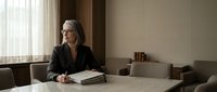Professional mature businesswoman with gray hair sitting at conference table in modern office interior during daytime meeting preparation.