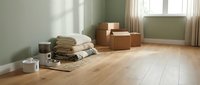 Empty living room with stacked cardboard moving boxes, folded textiles, and wooden flooring during relocation or home renovation project.
