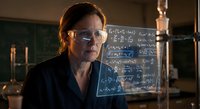 Middle-aged Caucasian female scientist in glasses working with mathematical equations on blackboard in laboratory setting for educational content.