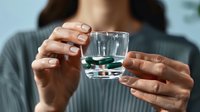 Woman holding glass of water and green capsule supplements, close up view of female hands with natural manicure showing daily vitamin intake routine.