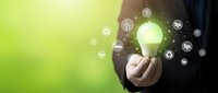 Businessman holding glowing lightbulb with renewable energy icons on green background representing sustainable innovation and eco-friendly technology solutions.