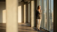 Young woman with brown hair, Caucasian ethnicity, wearing business casual attire with blazer and light pants, standing by large windows in modern office space with natural sunlight streaming through glass panels.