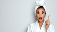 Young mixed race woman in white bathrobe and hair towel showing surprised expression with open mouth and raised finger against light background. Beauty and skincare concept.