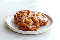 Freshly baked glazed French crullers with golden caramelized coating served on white ceramic plate against light background, showcasing flaky pastry texture.