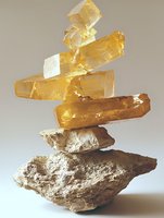 Stacked golden yellow crystals balanced on rough limestone rock formation against neutral background, showcasing natural mineral geometry and light interaction.