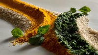 Vibrant lines of colorful spice powders with fresh basil leaves on light surface, dramatic side lighting creates texture and shadows in culinary composition.