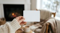 Caucasian woman holding blank white card in cozy living room with fireplace, perfect for holiday greetings, promotional messaging, or seasonal marketing campaigns.