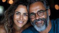Mature couple with genuine smiles, man with grey beard and glasses next to woman with wavy brown hair, close-up portrait against blurred bokeh lights background.