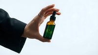 Hand holding green glass dropper bottle with amber liquid against white background for skincare cosmetics and wellness products.