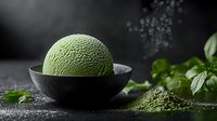 Green matcha ice cream scoop in black ceramic bowl with powdered tea and fresh mint leaves on dark background, moody food photography composition.