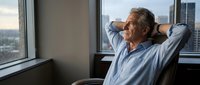Senior businessman relaxing in office chair with hands behind head, looking out window at city view. Corporate executive taking break from work.