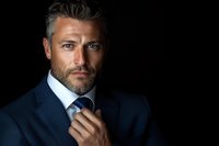 Confident mature businessman with gray hair and beard wearing navy suit and blue tie adjusts necktie against dark background in dramatic lighting.