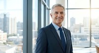 Confident mature Caucasian businessman in navy suit smiling in modern office with city skyline view through floor-to-ceiling windows during golden hour.