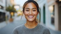 Young Asian woman with bright genuine smile wearing casual grey sweater outdoors in urban setting with soft natural lighting and blurred city background.