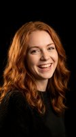 Happy young Caucasian woman with curly red hair smiling warmly in dark sweater against black background for portrait photography and lifestyle content.