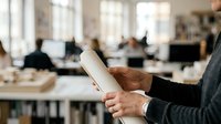 Architect or engineer holding rolled blueprint plans in modern office workspace with colleagues working in background.