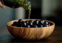 Fresh black olives in wooden bowl with pouring extra virgin olive oil, dark moody food photography on rustic table background for Mediterranean cuisine.