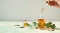 Hand holding dropper adding golden oil to glass bottle with eucalyptus leaves on white surface. Natural skincare, aromatherapy and wellness concept.