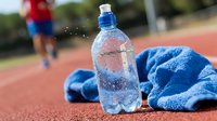 Bottle of water with condensation on athletic track next to blue towel, runner in background during outdoor fitness training session.